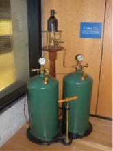 Photograph shows green tanks of the oil lamp, large pumping handle sits in front of the tanks. Small, black kerosene burner sits above the tanks and handle on a wooden pedestal.