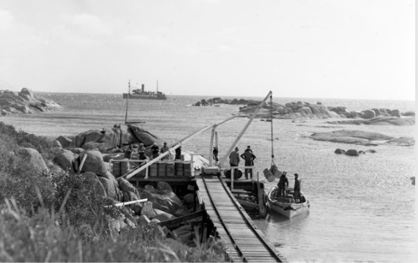 Black and white photograph showing a tramway beside a shoreline. A pier at the end of the tramway is filled with seven people, and a crane is lifting barrels into a small boat in the water below. Two people are standing in the boat helping load the cargo. A large ship can be seen in the distance. 