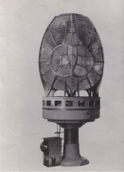 Black and white photograph showing large lighthouse lens and pedestal. Lens consists of many prisms arranged in concentric circles, round pedestal sits underneath with a small cranking mechanism on its left-hand side.