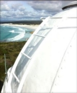 Photograph showing white ladder attached to the side of lantern roof dome. Beach and be seen in background. 