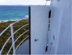 Photograph showing lantern room door on lighthouse balcony. White door with small handle is standing ajar. Balcony handrail can be seen behind door with view of sea beyond. 