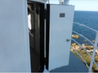 Photograph showing lantern house door. White door with small plaque and handle standing ajar onto balcony. Lantern room inside can be seen through opening. 