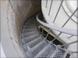 Photograph showing grey, iron, spiral stairs and railing leading down from lantern house. Staircase is embedded into granite wall on lefthand side.  