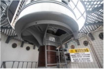 Photograph showing large grey lens pedestal within lantern room. Pedestal is supported on pillar with a small grey ladder attached at its base. Mesh security gate fixed to right-hand side of pedestal has large yellow and white 'Warning' sign attached. 