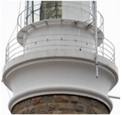 Photograph showing lantern house base including balcony balustrades, external catwalk and lower glass glazing panes. The top granite stones of the main tower can be seen below the base. 