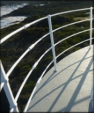 Photograph showing curved, white balcony stanchions and railings embedded in the grey balcony floor. Green vegetation can be seen on the ground below the lighthouse. 