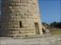 Photograph showing base of lighthouse tower. The light brown granite stones are arranged in a circle, and a window and door opening can be seen leading into the entrance floor. A grey concrete aprons runs around the perimeter of the tower, and a granite staircase can be seen around the corner of the tower. 