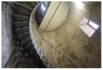 Photograph showing internal, light brown granite walls of the lighthouse tower. The camera points up inside the tower, and the cast iron stairs spirals its way up the wall, and it disappears onto an iron half-floor in the shape of a semi-circle above. 