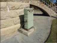 Photograph showing green utility box standing upright beside the base of the light brown tower. It is embedded in the concrete apron, and a granite flying staircase can be seen in the background. 