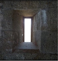 Photograph showing thin rectangular opening between granite stones. The window ledge is built into the stone, and bright sunlight can be seen through the opening.