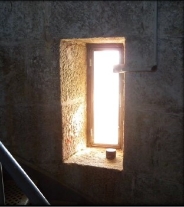 Photograph showing thin rectangular opening between granite stones. The window ledge is built into the stone, and bright sunlight can be seen through the opening.