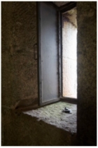 Photograph showing thin rectangular opening between granite stones. The window ledge is built into the stone, and bright sunlight can be seen through the opening. A door for the window is standing ajar. 