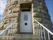 Photograph showing front door of lighthouse tower. A white, lined door embedded in the rough hewn granite stones of the tower at the top of a staircase which is obscured from view. A window can be seen above the door, and two signs are nailed to the front of the door. 