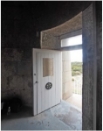 Photograph showing front door of lighthouse tower from the inside. A white, lined door embedded in the smooth granite stones and standing ajar. A window can be seen above the door, a sign is nailed to the front of the door. A round vent is nailed to the door.  