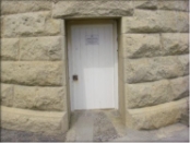 Photograph showing white entrance door at the base of lighthouse tower. A white, lined door embedded in the rough hewn granite stones of the tower. A sign is nailed to the front of the door. 