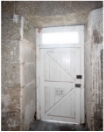 Photograph showing front door of lighthouse tower from inside. A white, lined door embedded in the smooth granite stones of the tower. A window can be seen above the door, and a bright light can be seen through the window. 