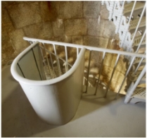 Photograph showing half-floor of tower. A railing and stachions are embedded in the floor, and a lattice staircase can be seen behind.  There is an arched iron cover that extends upright from the floor to meet the railing height, and is embedded in the railing. 