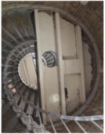 Photograph showing underside of semi-circle half-floor within lighthouse tower. The floor consists of iron sheeting rolled on beams and painted in a cream colour. The floor meets the internal wall on its arched side, and a grey lattice staircase spirals up the wall to meet the floor. A large hole in the floor shows a peephole on the levels above.  