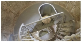 Photograph showing half-floor of tower. A railing and stanchions are embedded in the floor, and a lattice staircase is embedded into the half floor from the level below. Another lattice staircase starts on the opposite end of the semi-circle and advances to a higher level.  There is an arched iron cover that extends upright from the floor to meet the railing height, and is embedded in the railing. The cover is arched around a hole in the floor. Lower levels of the lighthouse can be seen below.  