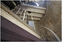 Photograph showing underside of two semi-circle half-floors within lighthouse tower. The floor consists of iron sheeting rolled on beams and painted in a cream colour. The floors meet the internal wall on their arched sides, and a grey lattice staircase spirals up the wall to meet the floors. A large hole in the floor shows a peephole on the levels above.  