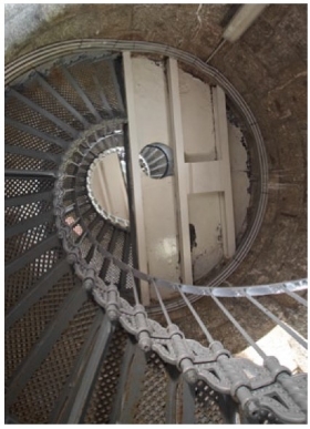 Photograph showing underside of semi-circle half-floor within lighthouse tower. The floor consists of iron sheeting rolled on beams and painted in a cream colour. The floor meets the internal wall on its arched side, and a grey lattice staircase spirals up the wall to meet the floor. A large hole in the floor shows a peephole on the levels above.  