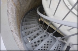 Photograph showing grey lattice stairs. The steps are embedded in the wall and wraps around and down to a lower level. Wrought iron handrails are attached to the inside of the stairs from the base of each step. 