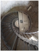 Photograph showing underside of semi-circle half-floor within lighthouse tower. The floor consists of iron sheeting rolled on beams and painted in a cream colour. The floor meets the internal wall on its arched side, and a grey lattice staircase spirals up the wall to meet the floor. A large hole in the floor shows a peephole on the levels above.  