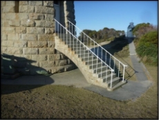 Photograph showing external flying staircase. The staircase extends from the upper entrance level of the rough-hewn granite tower down to the concrete apron below. White, thin handrails and stanchions are embedded in each step. A track leads from the base of the stairs into the vegetation where a small white domed building can be seen. 