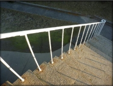 Photograph showing thin, white staircase railing. Each stanchion is embedded in granite steps leading down to a concrete base on the ground level. 