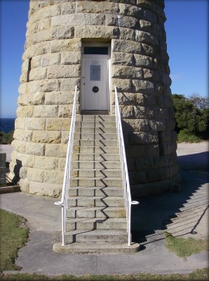 Photograph showing front door of lighthouse tower. A white, lined door embedded in the rough hewn granite stones of the tower at the top of a staircase which leads down to a concrete surface at the base of the lighthouse tower. A window can be seen above the door, and two signs are nailed to the front of the door. 