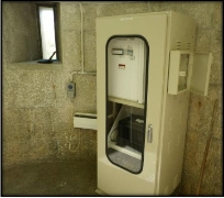 Photograph showing an upright utility box beside a smooth granite stone wall. The box has a clear window on its front, showing equipment on the inside. 