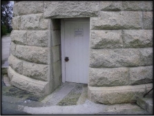 Photograph showing white entrance door at the base of lighthouse tower. A white, lined door embedded in the rough hewn granite stones of the tower. A sign is nailed to the front of the door. 