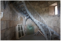 Photograph showing grey lattice staircase spiralling up a curved granite stone wall. A window opening is cut into the stone and a bright light shines through from the outside. Glazing panes and frames sit piled together on the left side of the room on the ground.  