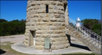 Photograph showing base of lighthouse tower. The light brown granite stones are arranged in a circle, and a window and door opening can be seen leading into the entrance floor. A grey concrete aprons runs around the perimeter of the tower, and a granite staircase can be seen around the corner of the tower. 