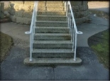 Photograph showing granite stairs with thin white handrail stanchions leading down to a concrete base. 