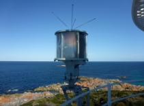 Photograph showing round, modern beacon with four antenna protruding from top. It is attached to a white handrail, and the rocky shoreline can be seen in the background below. 