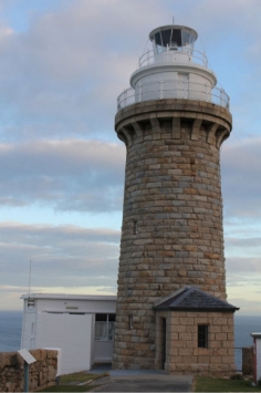 Photograph showing granite lighthouse tower. The tower is constructed of rough-hewn stone, and a white lantern house sites atop. A small porch with a black roof is attached to the front of the tower. A white building of modern original is half-concealed behind the tower. 