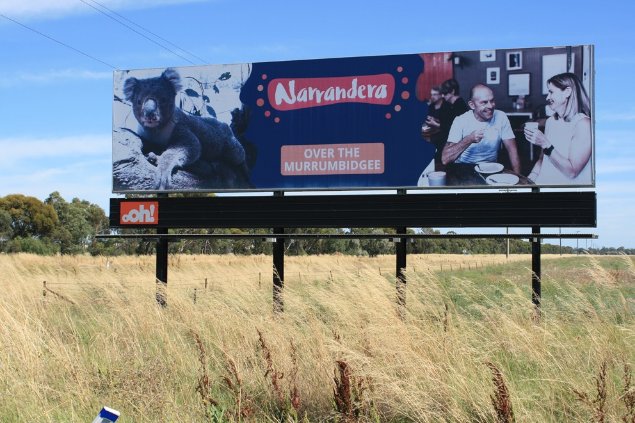 Signage advertising the local Koala attraction, welcoming visitors. Narrandera, central NSW. 
Image: © S. Brown.  

Photograph of a large landscape advertising sign along a roadside with grassy understory, open blue sky. The sign has images of a koala on the left hand side; on the right-hand side an image of a man and woman enjoying a coffee in a café and the words Narrandera, over the Murrumbidgee.  