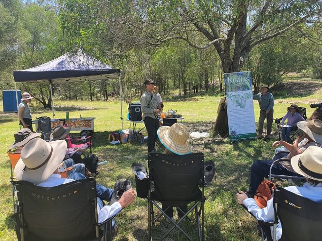 A community group receiving instructions on surveying Koalas, Gympie, Qld. Image: © CSIRO.

Photograph of a group of people, seated in deck chairs, under the shade of a tree facing a person giving instructions. A large billboard has information on koalas.  