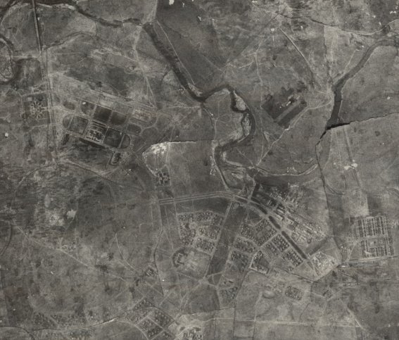 Black-and-white photograph with a river to the top and indications of road layouts, landscaped trees and buildings below.  Shows a triangular arrangement of roads to the top left.