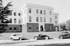 Black-and-white photograph of three-storey building with a central square doorway.  Three cars are parked in the foreground.
