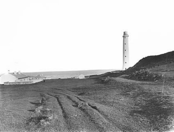 A black and white photograph showing a tall, white lighthouse in an open landscape with a collection of houses with chimneys to the left of the tower.  