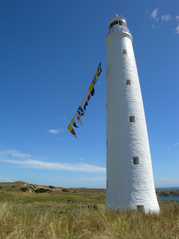 Photograph showing a tall, white lighthouse tower against a blue sky with ten flags of a variety of shapes and patterns attached to the base of the tower's balcony.