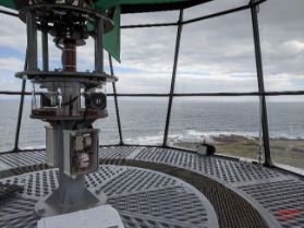 Photograph showing inside view of glazing panes with cast iron lattice floor. Mechanical pedestal stand is bolted to centre of lattice floor. Grey sea can be seen through glazing panes outside. 