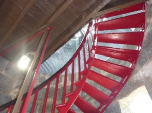 Photograph showing the underside of a red spiral staircase leading to a timber floor above. 