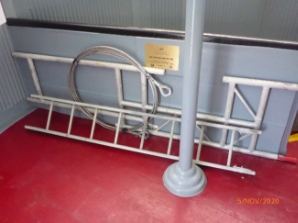 Photograph showing red timber floor with a thin white ladder leaning against a grey half-wall (amongst other structural equipment. A plaque can bee seen fixed to the half-wall, and a cast iron post is fixed to the red floor. 