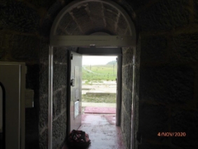 Photograph showing arched frame door way. Open door leads out to grass outside.