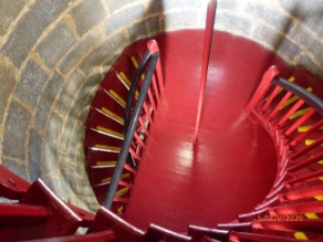 Photograph showing aerial view of round, internal lighthouse tower landing. Timber floor is painted red, and a red spiral staircase wraps around grey tower walls and descends to floor below. A red spiral staircase wraps around grey tower walls and ascends to floor above. 