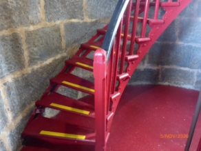 Photograph showing red spiral staircase fixed to curved stone wall. The staircase has black timber handrails and each tread has a strip of yellow at edge.