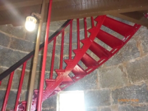 Photograph showing underside of red spiral staircase ascending to timber floor above. Staircase is fixed to curved, grey stone wall and has a black handrail.